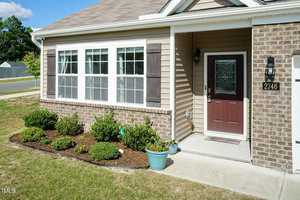 Front view of a house with tan siding and brick accents. It features a dark wooden door with a glass panel, a row of windows, and neatly trimmed shrubs in a small garden along the sidewalk.