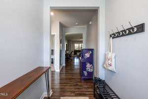 A modern hallway with wooden floors, a coat rack with a bag hanging, and a bench with shoes. A purple cabinet with floral designs stands on the right, leading to a living area visible in the background.