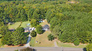 Aerial view of a rural property surrounded by dense trees. The area features several buildings, a circular pool, and an open grassy field. A narrow road runs along the bottom of the image, with driveways leading to the houses.