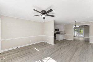 A bright, empty living space with light wooden floors and neutral walls. It features a ceiling fan and an open layout leading to a kitchen area in the background. French doors with windows open to an outdoor area.