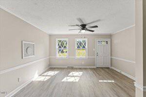 A bright, empty living room with light-colored walls and wood flooring. It features two windows with crossbar patterns, a ceiling fan, and a white door. Sunlight casts geometric shadows on the floor.