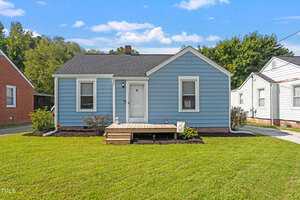 A small blue house with white trim sits on a neatly manicured lawn at 310 E Church Street, Creedmoor. It features a simple wooden porch and a white door. The sky is clear with a few clouds, and neighboring houses are partially visible.