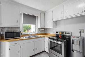 A small kitchen with white cabinets, a stainless steel stove, and a microwave. There's a double sink under a window with a gray blind. A gray backsplash and a compact water dispenser are also visible.