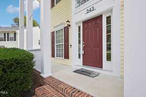 The image shows the entrance of a house with a maroon front door and white trim, accented by a black welcome mat. The house features yellow siding and white columns, with a small brick step leading to the porch. A hedge borders the walkway.