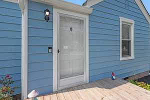 A small blue house with a wooden deck entrance, featuring a white door and screen door. There is a lantern-style light fixture beside the door and a decorative garden gnome on the deck. A window is visible to the right of the door.