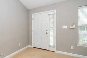 A gray-walled room featuring a white door with an adjacent window covered by white blinds. There's a security keypad on the wall beside the window, and the floor is carpeted in beige.