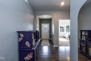 A hallway with wood flooring features a floral-patterned cabinet on the left. A front door with frosted glass is visible in the distance, and partially open double doors on the right reveal a lit room with a window.