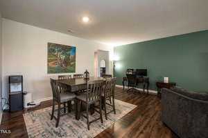 A cozy living space with a dark wooden dining table and six chairs on a patterned rug. A computer desk with a monitor and lamp is against a green accent wall. A colorful painting hangs on a light wall. The floor is polished wood.