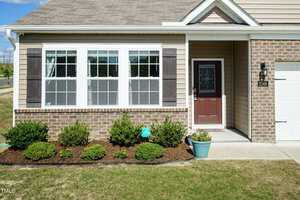 A suburban home featuring a brick facade and beige siding. Large windows with shutters are at the front, along with a maroon front door. Small shrubs and potted plants decorate the neatly maintained lawn and garden bed.
