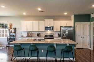 A modern kitchen featuring white cabinets, a stainless steel oven, microwave, and refrigerator, with a large granite island and four green velvet barstools. The room has wooden flooring and ample lighting.