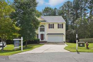 A yellow two-story house with a black garage door and white trim, located on a cul-de-sac. It has a concrete driveway, a mailbox near the road, and trees surrounding the property. A "For Sale" sign is visible on the left of the driveway.
