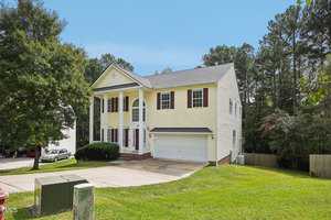 A two-story yellow house with white columns and red shutters is situated in a grassy yard. It features a double garage, a gabled roof, and a driveway. The background displays a row of trees and a bright blue sky.