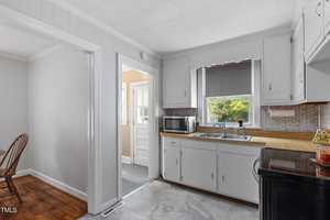A bright kitchen with white cabinets, wooden countertops, and a double sink under a window with a gray shade. A microwave sits to the left, and a door leads outside. Hardwood flooring is visible in the adjoining room.