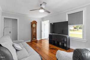 A living room with gray walls featuring a sofa and armchair, a large flat-screen TV on a black stand, and a wooden grandfather clock. A ceiling fan is on the white ceiling, and natural light comes through a window with a gray blind.