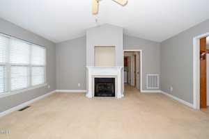A spacious, empty living room features a vaulted ceiling and a white fireplace centered on a taupe wall. Large windows with blinds provide natural light from the left, and there are doorways on either side of the fireplace. The floor is carpeted in beige.