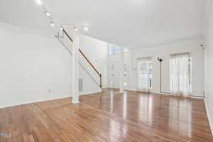Spacious empty living room with polished hardwood floors and tall white walls. Large windows with sheer curtains allow natural light to fill the space. A staircase with wooden railing leads to the upper level. Ceiling spotlights illuminate the area.