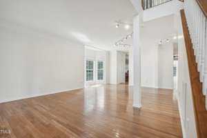 A spacious, empty living room with polished hardwood floors and white walls. Sunlight streams in through large windows. A staircase with a wooden handrail is on the right, and track lighting is on the ceiling.