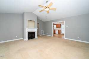 Spacious living room with gray walls and beige carpet. Features a white fireplace with a mantel and a ceiling fan. An open doorway leads to a bright kitchen with wooden cabinets and a window, visible in the background.