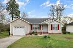 Nestled at 708 Ascot Drive, Creedmoor, this charming single-story house features beige siding, a gray roof, and red shutters. It comes with a one-car garage and a quaint front porch, surrounded by lush grass and a few trees under a partly cloudy sky.