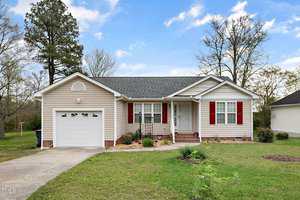 Nestled at 708 Ascot Drive, Creedmoor, this charming single-story house features beige siding, a gray roof, and red shutters. It comes with a one-car garage and a quaint front porch, surrounded by lush grass and a few trees under a partly cloudy sky.