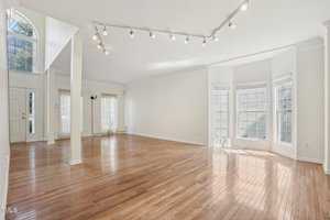 Spacious living room with hardwood floors, large windows, and white walls. Ceiling track lighting and a high, arched entrance with additional windows bring in natural light. Open floor plan leading to other areas.