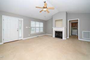 A spacious, unfurnished living room with beige carpet and light gray walls. It features a ceiling fan, a white fireplace, a door leading outside, and an adjacent hallway leading to a laundry area with visible appliances. Natural light comes from a large window.