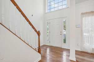 Bright entryway featuring a wooden staircase with white balusters. The space has a wooden floor and high ceiling, with a large window and a white front door flanked by narrow glass panels. Natural light streams in, creating a welcoming atmosphere.