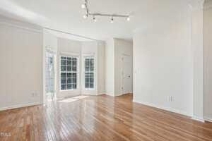 A bright, empty room with large windows and wooden floors. The walls are white, and there is a door in the corner. Ceiling lights are positioned above, creating a clean, open space with abundant natural light.