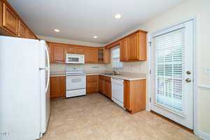 A kitchen with wooden cabinets, a white refrigerator, stove, and dishwasher. It has a beige tile floor, white countertops, and a window with blinds above the sink. A glass door with blinds leads outside, and recessed lighting brightens the room.