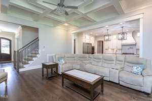 A spacious, modern living room with a coffered ceiling and wood flooring. It features a large beige sectional sofa, wooden coffee and side tables, and an open view of a white kitchen with pendant lights. A staircase is visible on the left.