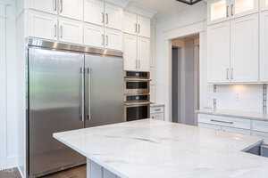 A modern kitchen featuring a large stainless steel refrigerator, double wall ovens, white cabinetry, and a spacious marble island countertop. The room is bright with ample natural light.