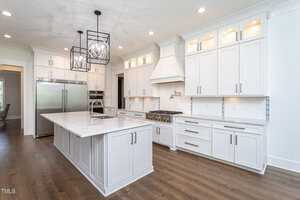 Modern kitchen with white cabinets, island with marble countertop and sink, stainless steel appliances, and dark wood flooring. Pendant lights hang above the island. The room is spacious and bright.