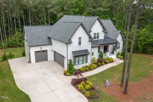 The two-story white house at 675 Willard Drive in Creedmoor boasts a gray roof and is surrounded by trees. With black window accents, it offers a spacious driveway to a two-car garage, complemented by well-maintained landscaping featuring green shrubs and a small stone garden.