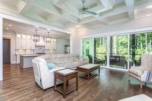 Spacious living room with coffered ceiling, light-colored sofas, and a wooden coffee table on polished wood floors. Large glass doors open to a balcony with forest views. Adjacent to an open kitchen with white cabinetry and hanging lights.