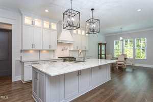 A modern kitchen with white cabinets, a large marble island, two geometric pendant lights, and a sink. Hardwood floors extend to a dining area with a table and chairs near large windows. A glass cabinet stands in the corner.