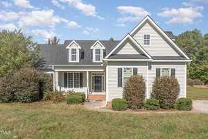 The white house at 104 Keeneland Drive boasts a gray roof, charming dormer windows, and a welcoming front porch with brick steps. Nestled in lush bushes and grass, this Oxford gem sits under a clear blue sky with scattered clouds.