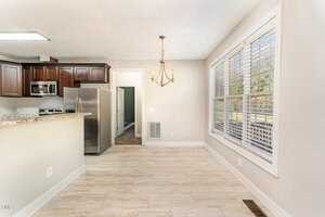 A bright kitchen with stainless steel appliances, dark wood cabinets, and a granite countertop. To the right is a dining area with a chandelier and large windows, allowing ample natural light. Light-colored tiled floors throughout.