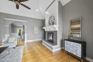 A modern living room with hardwood floors, a tiled fireplace, and a large clock above the mantel. It features a light gray armchair, a colorful abstract painting, and a dark dresser with decorative items. An arched doorway leads to another room.