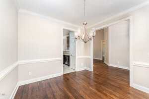A bright, empty dining room with hardwood floors and beige walls. A modern chandelier hangs from the ceiling. The room connects to a kitchen and hallway through open doorways.