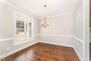 A bright, empty dining room with light gray walls, white trim, and hardwood floors. A chandelier hangs from the ceiling. Sunlight streams through a window with white blinds.