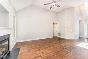 A spacious, empty living room with hardwood floors, a ceiling fan, and neutral-colored walls. A fireplace is in the corner, and a hallway leads to other rooms. Large windows allow natural light to fill the space.