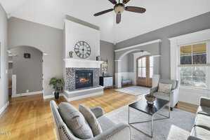 A spacious living room with a vaulted ceiling features a large clock above a modern fireplace. Two armchairs and a sofa surround a coffee table, with light wood flooring and arched doorways adding elegance. A ceiling fan is overhead.