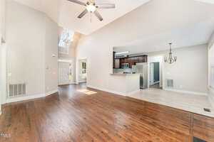 Spacious open-concept living area with wooden floors and vaulted ceilings. A ceiling fan hangs above the room. The kitchen is visible with stainless steel appliances, and a dining area with a chandelier is adjacent. Natural light streams through windows.