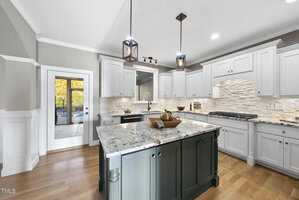 A modern kitchen with white cabinets, a large granite island with a dark base, and pendant lights overhead. Hardwood floors and a glass door leading to an outdoor area are visible.