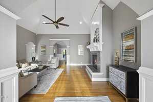 Nestled at 1107 Dovefield Lane in Youngsville, this spacious modern living room boasts high ceilings and hardwood floors. A cozy fireplace, neutral-toned furniture, abstract wall art, and a ceiling fan complement the space as light pours in from a distant window, enhancing the elegant atmosphere.
