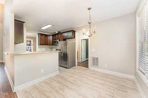 A bright kitchen with wooden cabinets, stainless steel appliances, and a granite countertop. An adjacent dining area features a chandelier and large windows. The floor is tiled in light beige, and the walls are painted a soft neutral color.