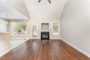 A spacious living room features a high ceiling, hardwood floors, and a central fireplace flanked by windows. Natural light fills the room from large windows and a glass door leading to an outdoor area.