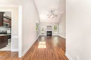 Bright living room with hardwood floors, a ceiling fan, and a fireplace. Large windows and glass doors let in natural light. To the left, an open doorway leads to a kitchen with dark cabinets and stainless steel appliances.