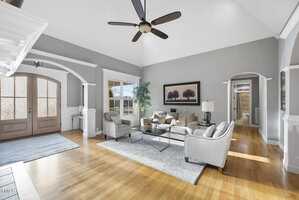 Spacious living room with high ceiling, large fan, and light wood flooring. Gray walls feature a picture and arched doorway leading to a hallway. Two sofas and a glass coffee table center the space, with natural light streaming through large windows.
