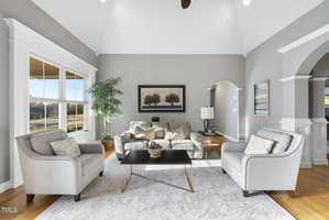A bright, elegant living room with high ceilings. It features a beige sofa, two gray armchairs, a modern coffee table, and a large window with views. The room has a neutral color palette and a framed nature painting on the wall.